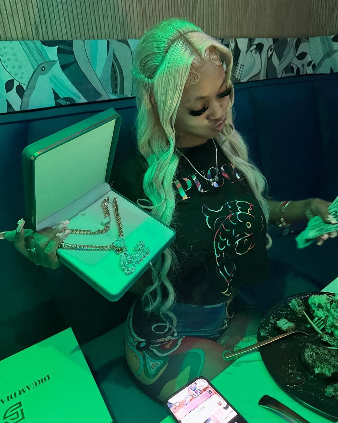 Bigg Britt with long blonde hair in a green-lit lounge, holding up an open jewelry box.