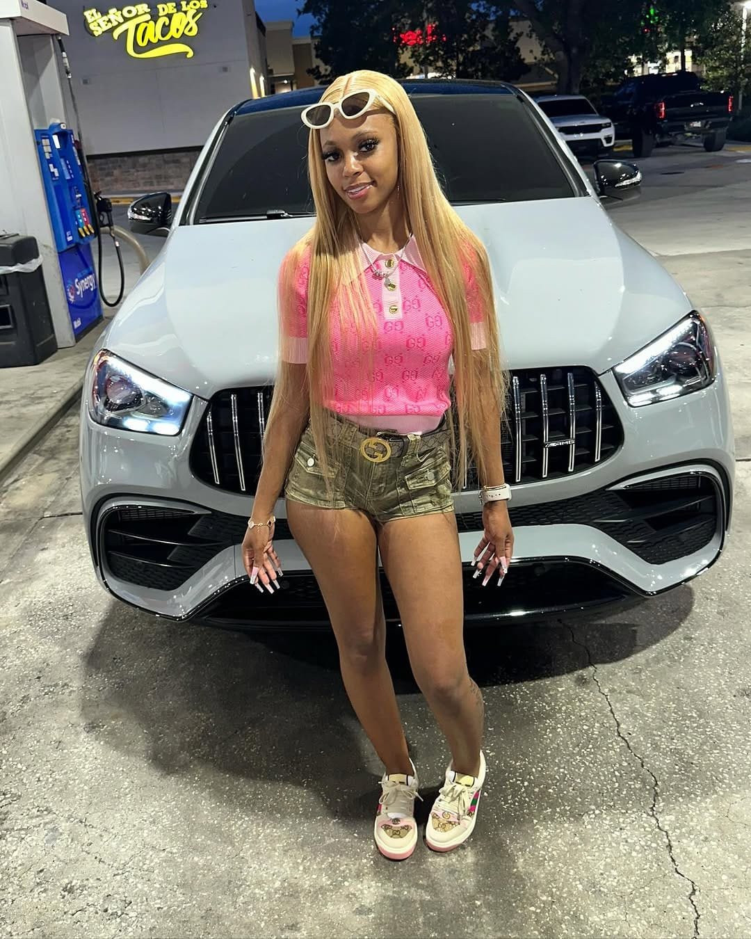Bigg Britt in a pink top, sunglasses pushed up on her long blonde hair, standing in front of a white Mercedes SUV.
