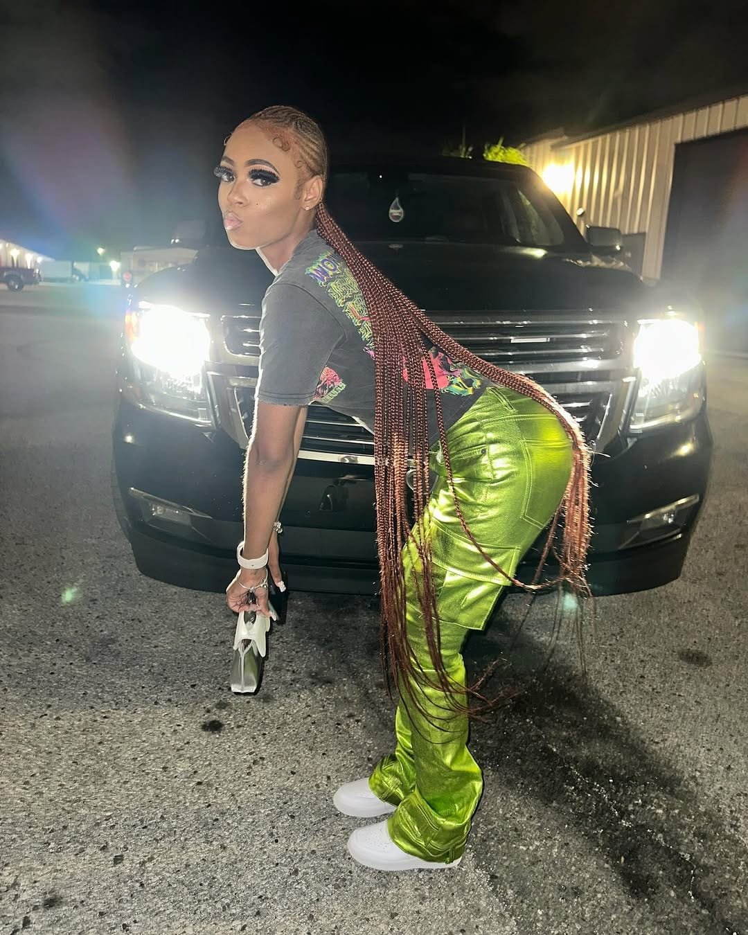 Bigg Britt with long braids, in a black top and green pants, posing beside a black SUV with its headlights on at night.